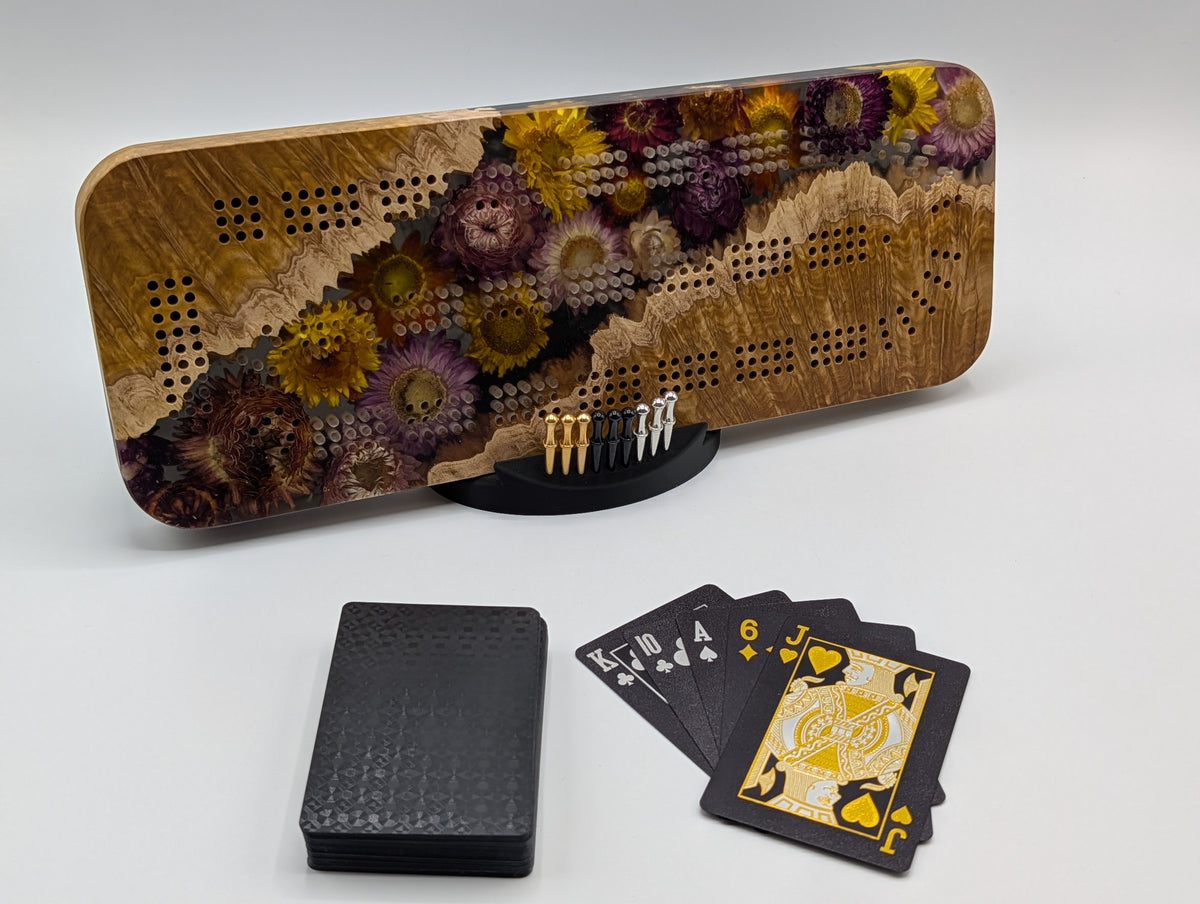 One-of-a-kind Imprint brown mallee burl cribbage board with real strawflowers in yellow, cream and purple, on its included display stand with pegs and a black playing-card deck.