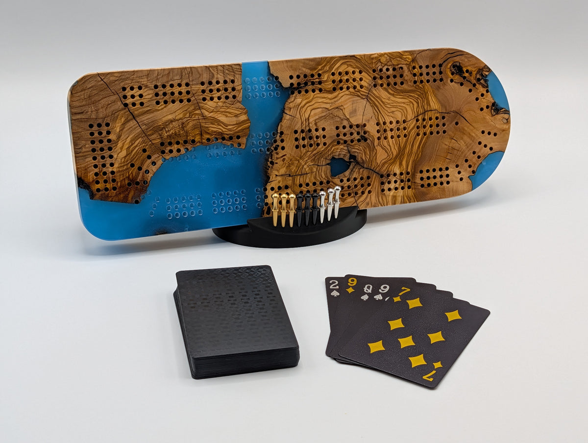 Azure Cove Origin Turkish olivewood cribbage board with bright blue resin — shown with included cards, pegs and display stand.