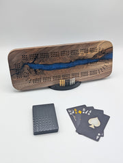 Origin Canadian black walnut cribbage board with a winding cobalt blue epoxy river, displayed with its cards, pegs and stand.