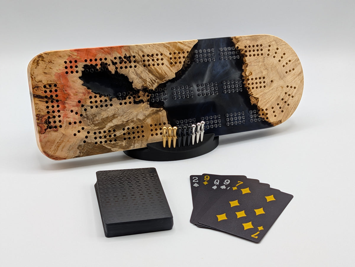 Ember Current Origin Manitoba maple burl cribbage board with smoky midnight blue resin — shown with included cards, pegs and display stand.